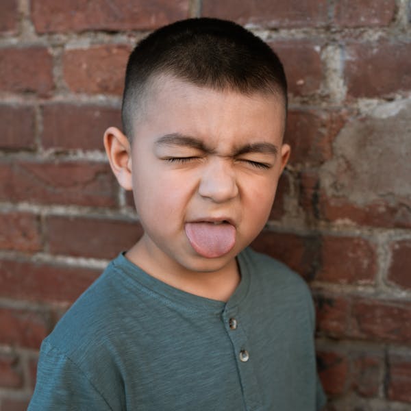 A cheerful boy in a gray shirt makes a funny face with closed eyes and sticking tongue out.