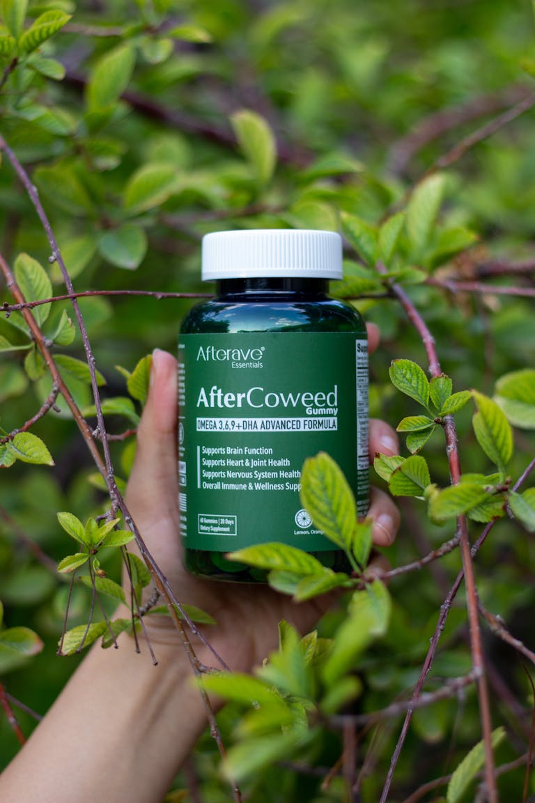 Hand holding a supplement bottle amidst vibrant green foliage outdoors.